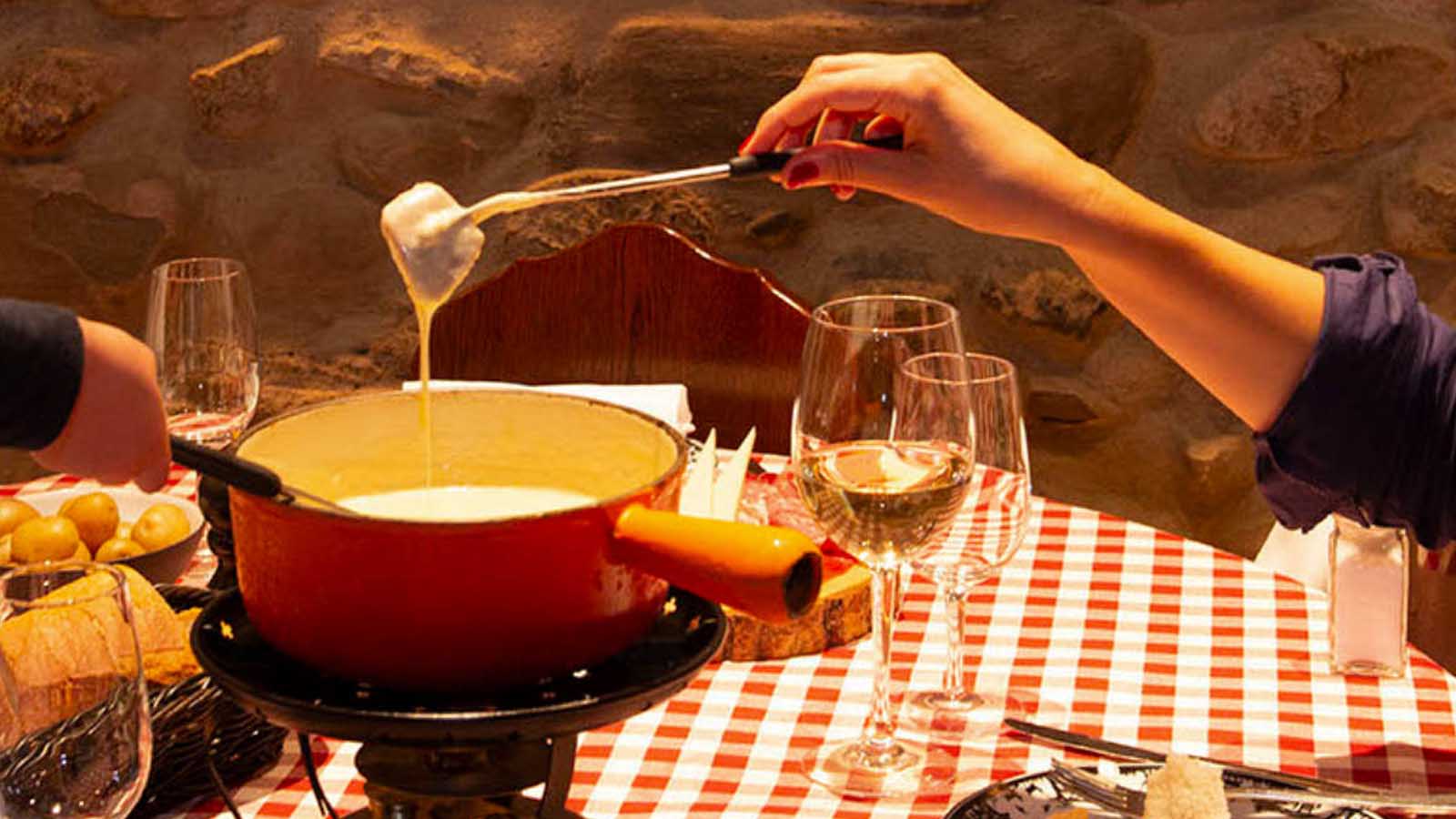 Hand holding a fork with bread over a red cheese fondue pan on a red and white checkmate tablecloth with a glass of white wine