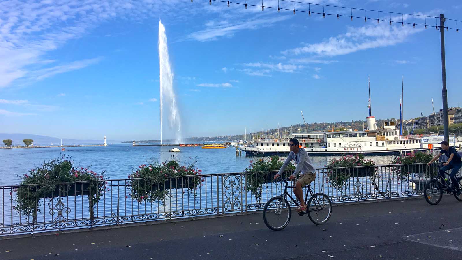 two men biking along the Geneva lake shore with the Geneva Jet d'eau (Geneva Fountain) and a white boat in the background